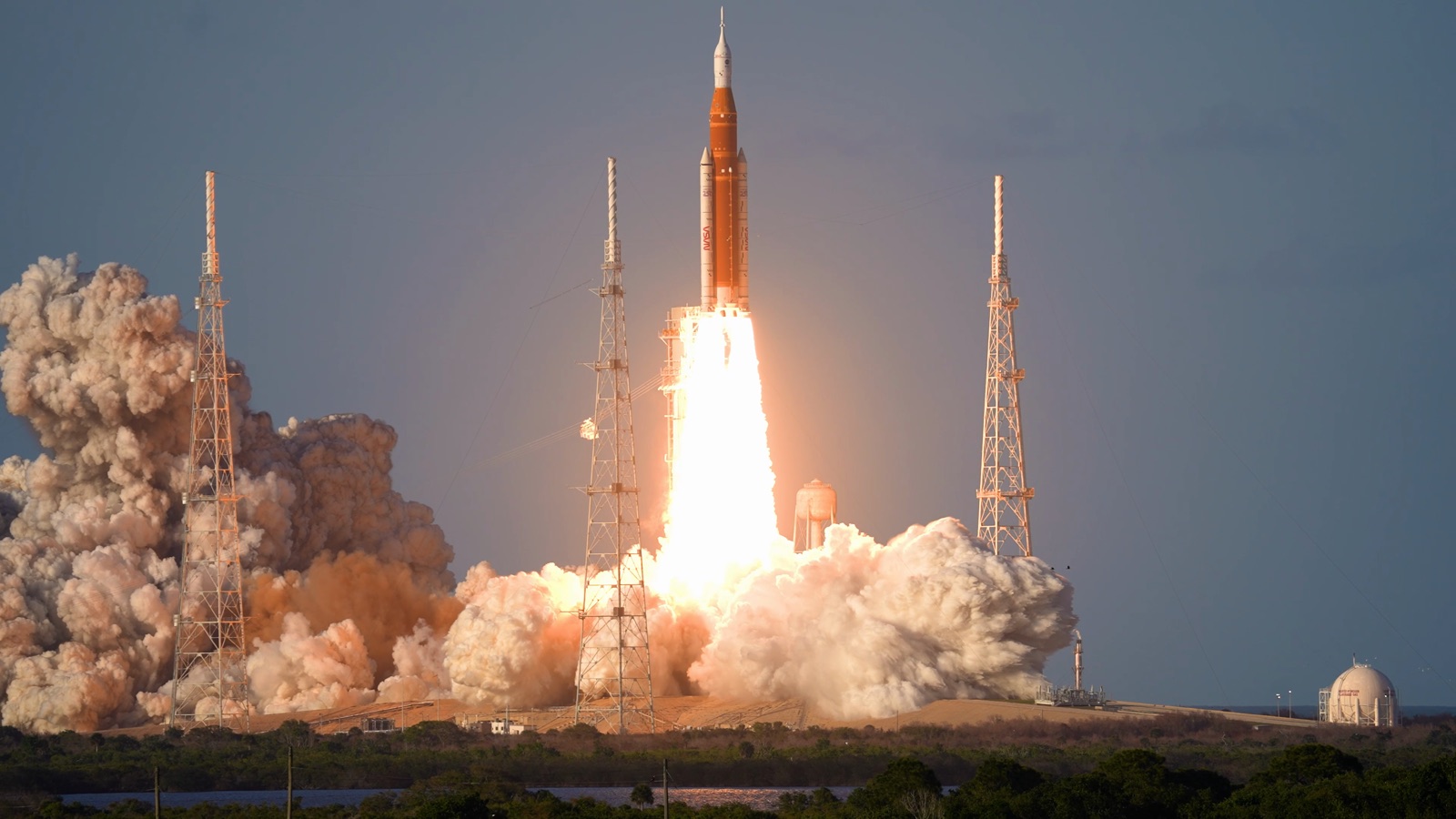 Artemis II's Space Launch System rocket rising past its launch tower on a column of fire, with clouds of exhaust billowing on both sides